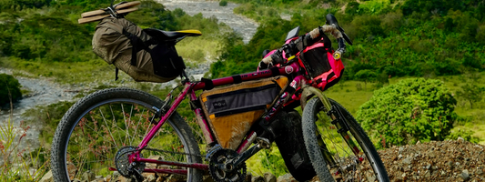 Ein pinkes Gravelbike mit mehreren Gepäcktaschen steht vor einer grünen, hügeligen Landschaft mit Fluss im Hintergrund.